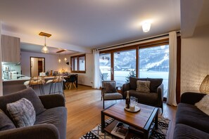 A modern living room with a sofa, armchair, and coffee table, a kitchen with a dining area, and large windows with a snowy mountain view. - Abela - Propriété d'exception, vue panoramique (Saint-Chaffrey)