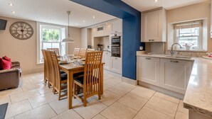 A modern kitchen with white cabinets, a dining area with wooden chairs, a clock on the wall, and a large window. - 4 Bed in Keswick (oc-b45231) (Keswick)