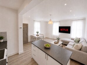 A modern kitchen with a central island, a dining area with a table and chairs, a living room with a sofa and a TV displaying 'NETFLIX', and a hallway with a door. - Luxury 4-Bed Apartment (Northolt)