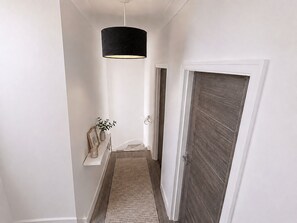 A hallway with a wooden door, a white wall, a black pendant light, and a small shelf with a plant and a picture frame. - Luxury 4-Bed Apartment (Northolt)