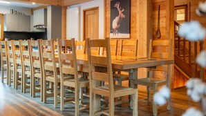 A dining area with wooden chairs and a table, wooden walls, a kitchen in the background, and a wooden staircase. - Le Stam - Seven Bedroom House, Sleeps 15 (La Salle-les-Alpes)