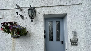 Pared blanca texturizada con una puerta azul, una cesta de flores colgante, un banco y un letrero que dice 'THREE TWO'.