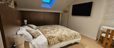 Un dormitorio con cama, mesita de noche, lámpara, televisor y tragaluz.