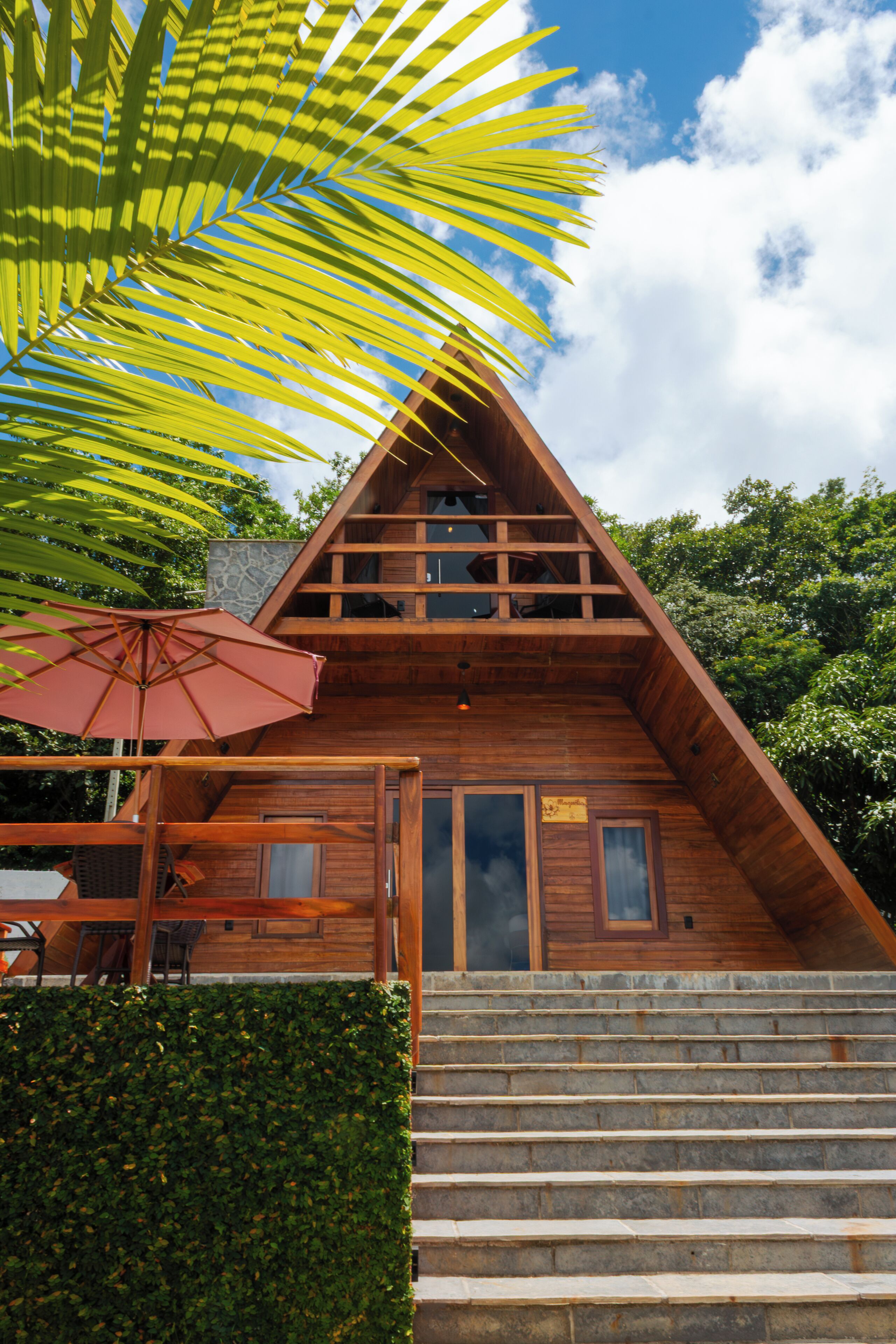A wooden cabin with a staircase leading to a balcony, surrounded by lush greenery.