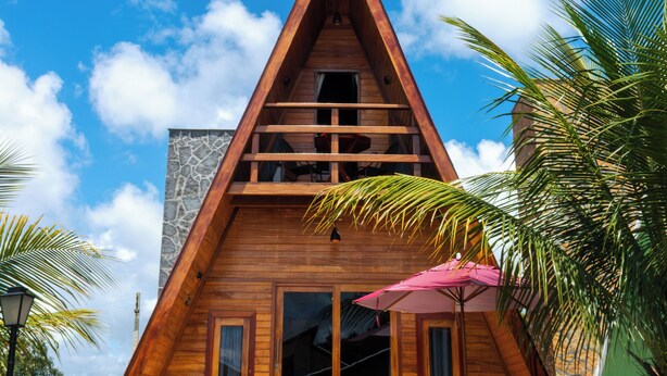 A wooden triangular building with a stone facade, a staircase leading to the entrance, and a patio area with a chair and umbrella.