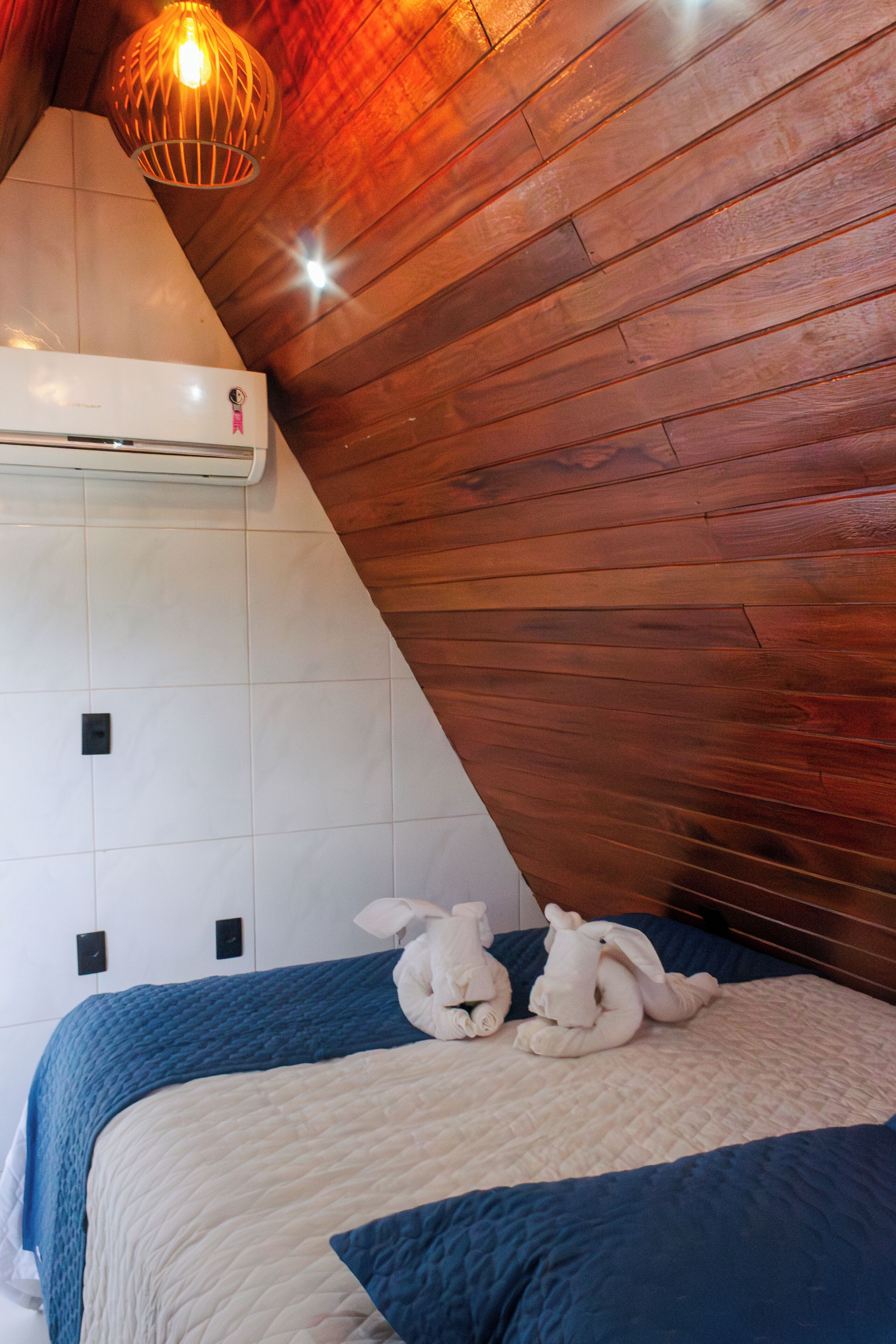 A bedroom with a sloped ceiling, wooden paneling, a bed with blue and white bedding, two plush animal pillows, and a hanging light fixture.