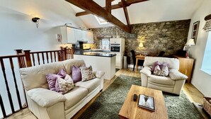A cozy living room with a sofa, armchair, and coffee table, a kitchen with stone walls, and a balcony with wooden beams. - 1 Bed in Keswick (oc-b45234) (Keswick)
