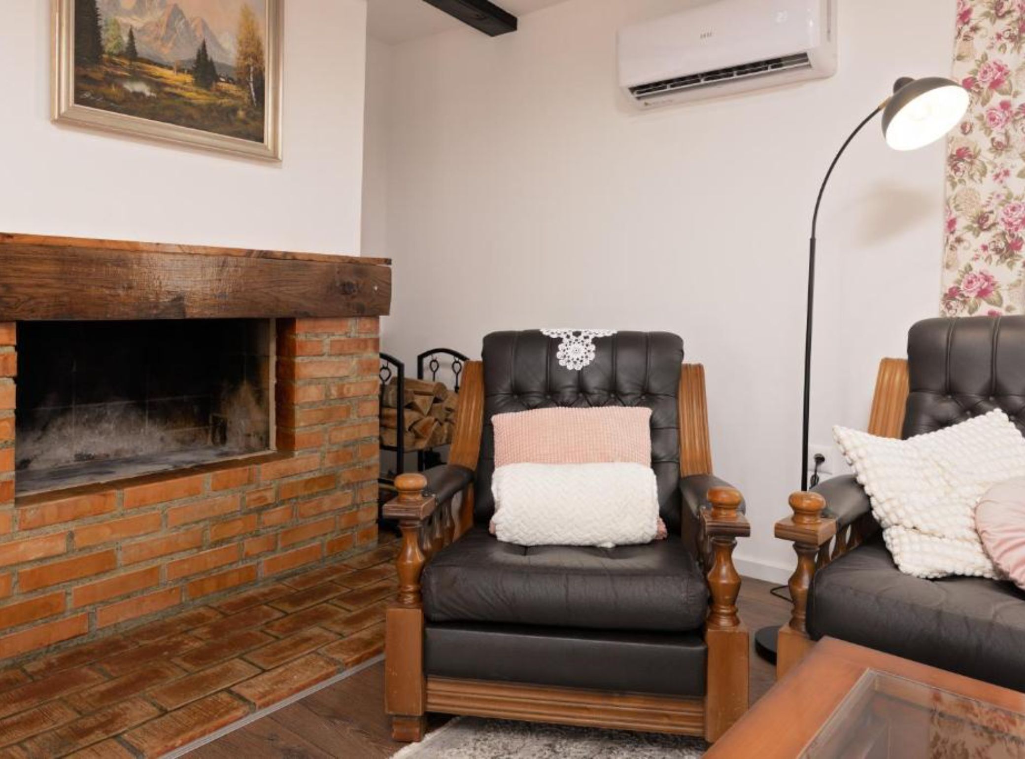 Una sala de estar con chimenea de ladrillo, un sillón de cuero negro, una mesa de centro de madera y una lámpara de pie.