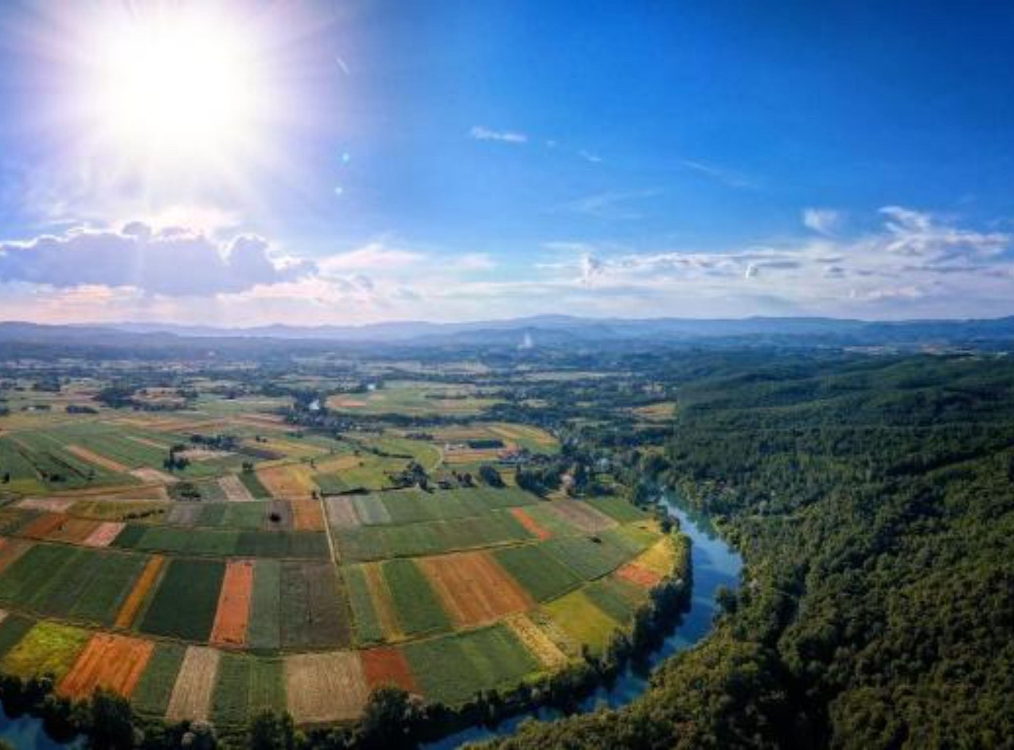 Vista panorámica de un paisaje rural con campos, un río y colinas boscosas.