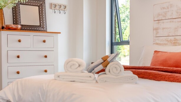 A neatly made bed with rolled towels and a red throw blanket, a white dresser with drawers, a mirror, and heart-shaped hooks on the wall. - 2 Bed in West Putford (oc-u45226) (Sessacott)