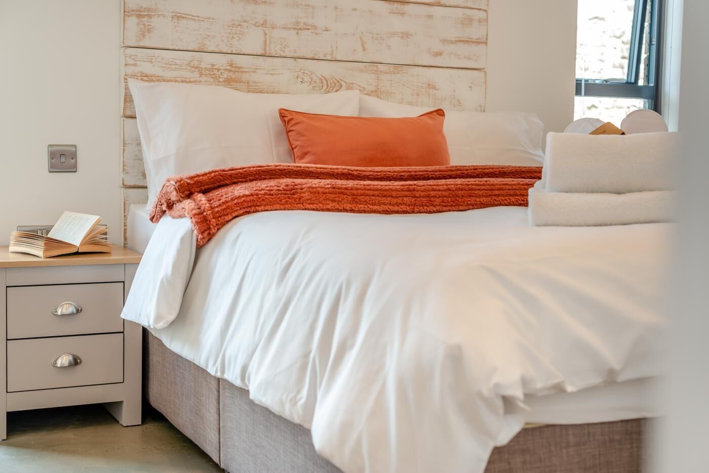 Un lit bien fait, avec une literie blanche et un seul oreiller orange, une table de chevet avec un livre, et un mur orné d’un panneau en bois au style rustique.