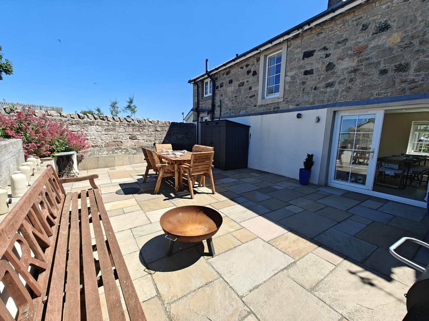 A patio with a wooden table and chairs, a barbecue grill, and a stone wall.