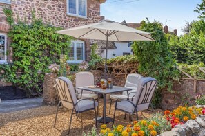 An outdoor patio with a table and chairs, surrounded by a stone wall and flowering plants. - 2 Bed in Carhampton (oc-v42711) (Carhampton)