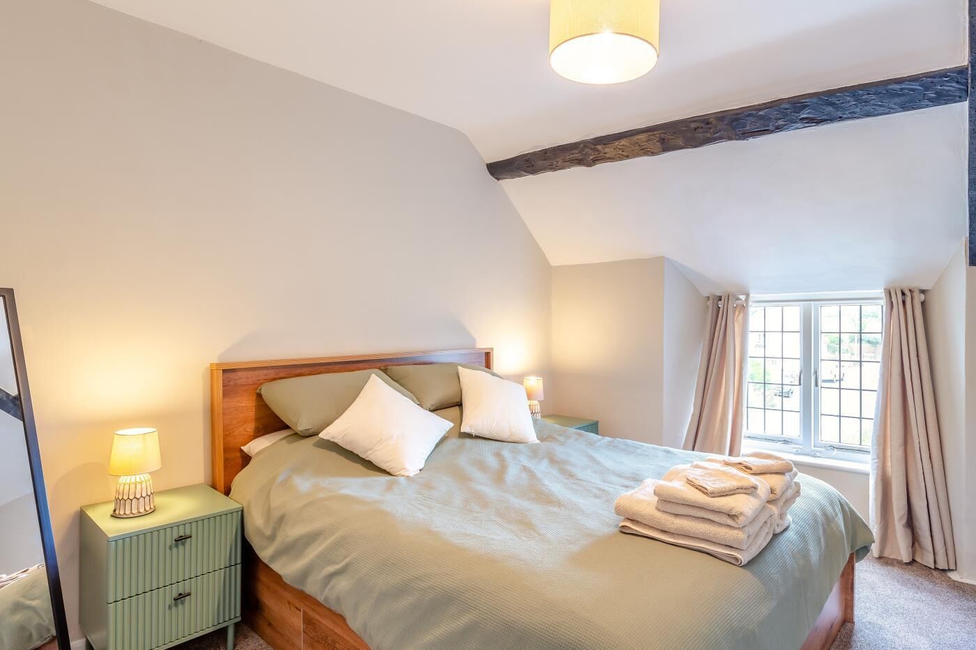 A bedroom with a bed, bedside tables, a window with curtains, and a wooden ceiling beam.