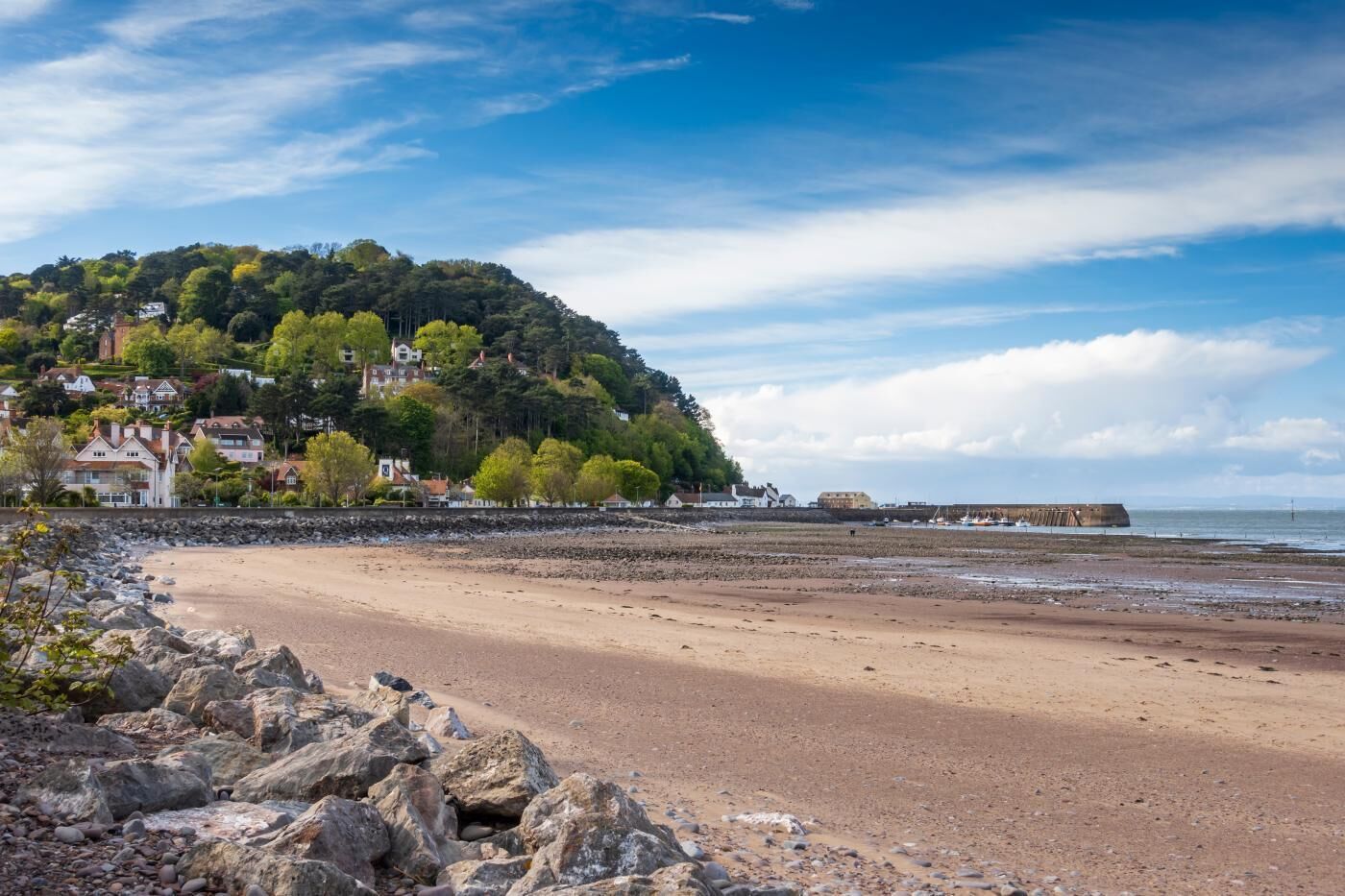 A coastal beach with rocky shoreline, a hill with houses, and a distant sea wall.