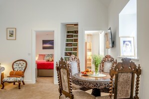 Ein Zimmer mit einem runden Holzes Esstisch, einem roten Bett, einem Bücherregal und einem gerahmten Bild an der Wand.