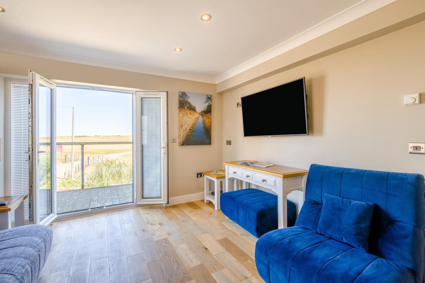 A room with a large window, a flat-screen TV mounted on the wall, a blue sofa, and a small table with a book.