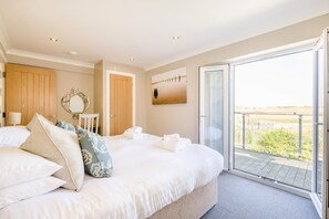 Un dormitorio con una cama grande, una puerta de madera, una silla blanca, una mesita pequeña y un balcón con vistas.