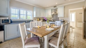 A kitchen with white cabinets, a wooden dining table, and chairs. There is a microwave on the counter, and a window with blinds. - 2 Bed in Old Hunstanton (oc-b44902) (Hunstanton)