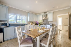 A kitchen with white cabinets, a wooden dining table, and chairs. There is a microwave on the counter, and a window with blinds. - 2 Bed in Old Hunstanton (oc-b44902) (Hunstanton)