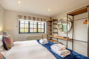A hotel room with a bed, a window with blinds, a radiator, a round mirror, and a built-in wardrobe with shelves. - 3 Bed in Hunstanton (oc-b44904) (Hunstanton)