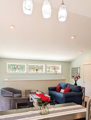 A living room with a blue sofa, a gray armchair, a coffee table, and a vase with red flowers. There are three pendant lights on the ceiling and a window with red flowers on the sill. - Peaceful Pines Ellsworth Apartment (Ellsworth)