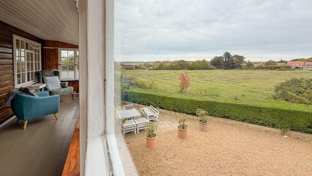 Ein Balkon mit Blick auf ein grünes Feld und einen Heckenstreifen, Holzwände und ein blauer Stuhl.