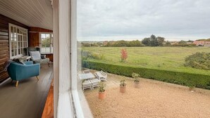 Ein Balkon mit Blick auf ein grünes Feld und einen Heckenstreifen, Holzwände und ein blauer Stuhl.