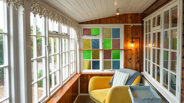 Ein gemütliches Zimmer mit einem gelben Sofa, einem kleinen Tisch und einem Erkerfenster mit bunten Glasfeldern.