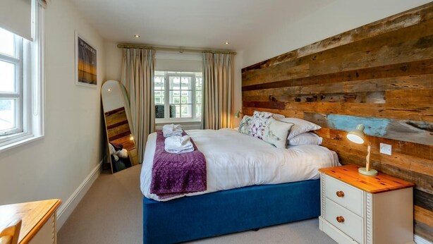 Quarto com cama, cabeceira de madeira, mesa de cabeceira com abajur, janela com cortinas e espelho.