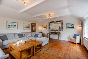 Ein Wohnzimmer mit einer Eckcouch, einem Holztisch, einem Konsolentisch mit Dekorationsobjekten, einem Spiegel und gerahmten Bildern an den Wänden.