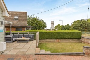 A residential backyard with a patio area, a grey sofa, a table with items on it, and a white tower in the background. - 4 Bed in Hunstanton (oc-o44907) (Hunstanton)