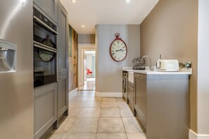 A modern kitchen with stainless steel appliances, a central island with a sink, and a wall-mounted clock. - 4 Bed in Old Hunstanton (oc-o44907) (Hunstanton)