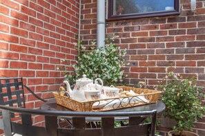 Ein Tisch mit Teekanne, Tassen und einem Brotkorb, an einer Ziegelwand mit Fenster und Grünpflanzen platziert.