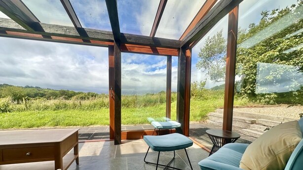 A modern outdoor patio with a glass ceiling, a wooden table, a blue cushioned chair, and a view of the countryside. - 2 Bed in Milton Damerel (oc-s42659) (Milton Damerel)
