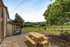 A patio with a wooden table and benches, a glass door leading to a room, and a vineyard in the background. - 2 Bed in Milton Damerel (oc-s42659) (Milton Damerel)