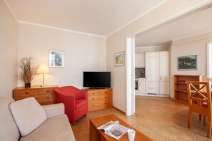 Un salon avec un canapé, un fauteuil rouge, une table basse en bois sur laquelle se trouvent un magazine et une tasse, un téléviseur posé sur un meuble en bois, et un coin cuisine à l’arrière-plan.