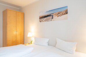 A bedroom with a wooden wardrobe, a bed with white bedding, a nightstand with a lamp, and a beach-themed wall art. - Strandnahe Ferienwohnung in Sellin mit Balkon (Sellin)