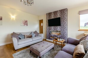 A living room with a grey sofa, a patterned ottoman, a fireplace, and a television mounted on a patterned wall. - 3 Bed in Dalwood (oc-c42675) (Dalwood)