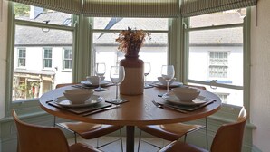 Een ronde eettafel met zes stoelen, een vaas met gedroogde bloemen en een uitzicht op gebouwen door grote ramen met gestreepte zonwering.