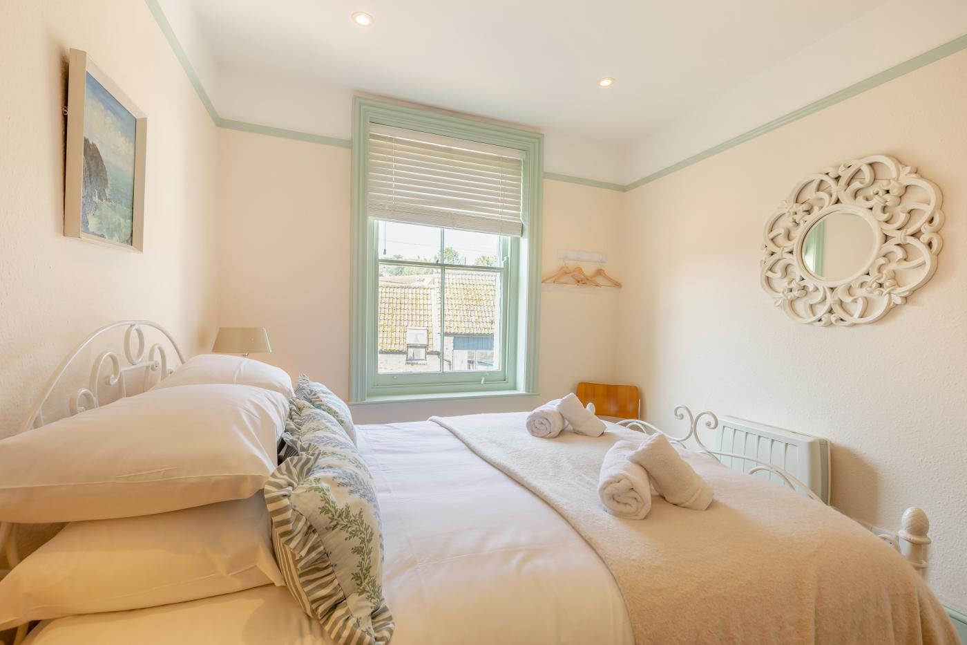 A bedroom with a double bed, a window with blinds, a bedside table, a wall-mounted mirror, and a painting on the wall.