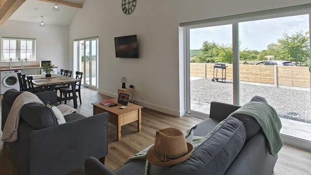 A spacious living area with a dining table, sofas, a TV, and a large sliding glass door leading to an outdoor area. - 3 Bed in Meidrim (oc-c45350) (Meidrim)
