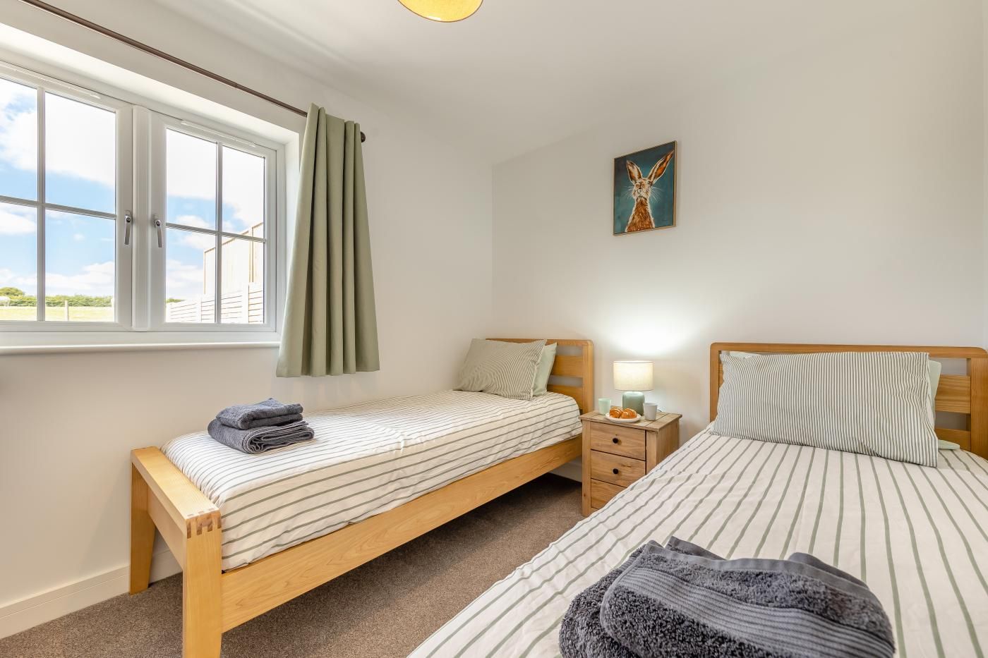 A bedroom with two single beds, a window with curtains, a bedside table with a lamp, and a painting on the wall.