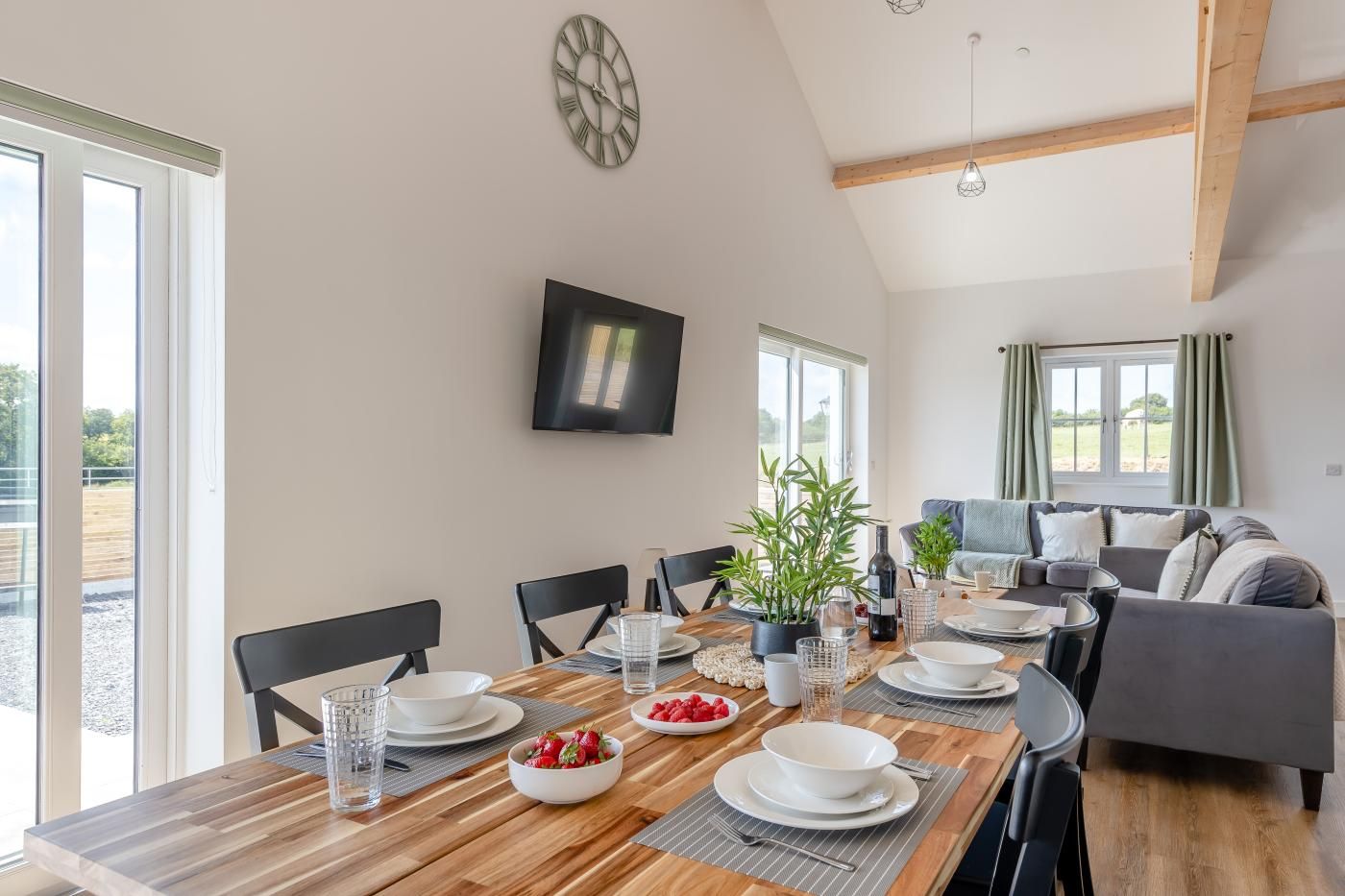 A dining area with a wooden table set for four, surrounded by chairs, with a television mounted on the wall above.