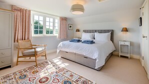 Een slaapkamer met een bed, een stoel, een nachtkastje, een lamp, een raam met gordijnen en een kamerkleed met patroon.