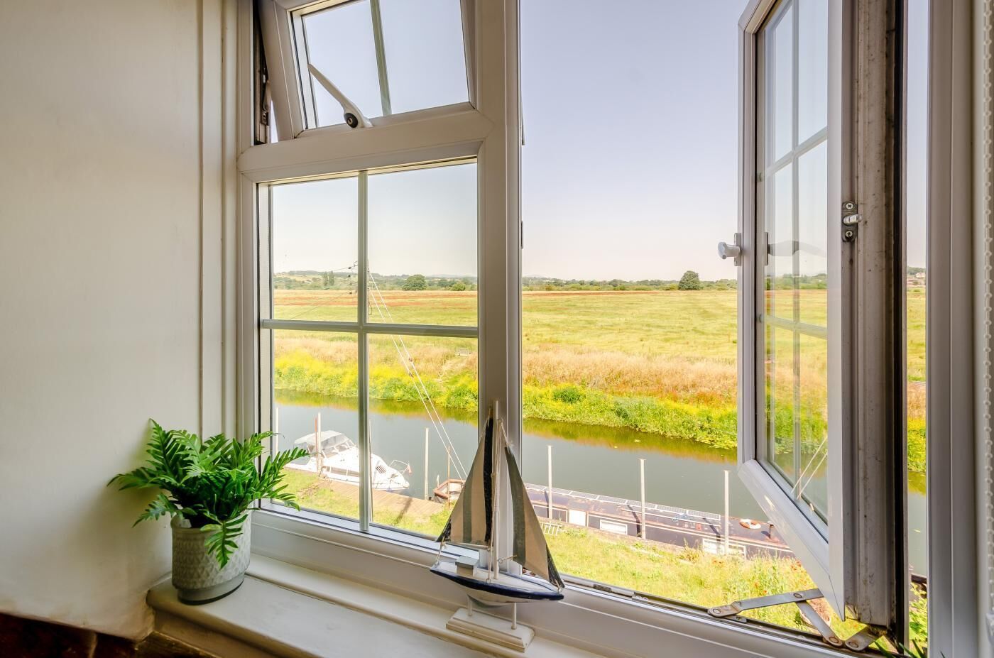 Ventana con vistas a un río y campos, una planta en el alféizar, y un modelo de velero.