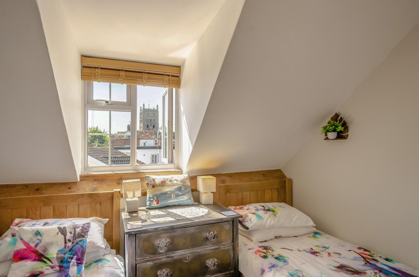 A bedroom with a sloped ceiling, a window with a view of buildings, a bed with a colorful duvet, a dresser with drawers, and a small plant on the wall.