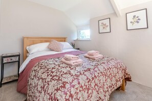 Une chambre à coucher comprenant un lit, une table de chevet, une fenêtre et deux œuvres d’art florales encadrées accrochées au mur.