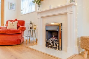 A living room with a fireplace, a red sofa, a small table with a bottle and glasses, and a wicker ottoman. - 4 Bed in Southwold (oc-t42835) (Southwold)