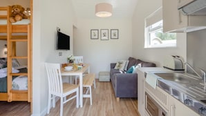 Un espacio habitable compacto con una cama litera, una zona de comedor con mesa y sillas, un sofá, una cocina con fregadero y estufa, y un televisor montado en la pared.
